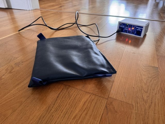 The Biomag 2-e therapy mat lying flat on a wooden floor, its lead running to the small control unit with its row of lit indicator lights.